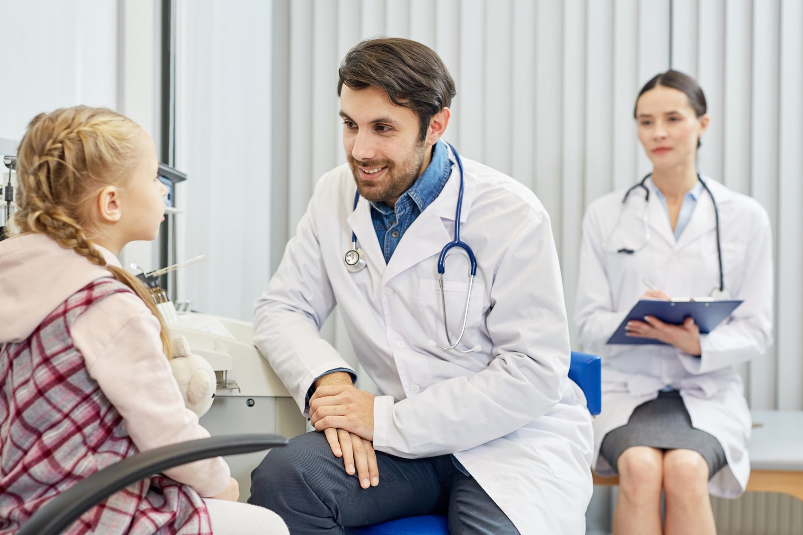 A doctor is speaking with a young girl while another healthcare professional takes notes in the background.