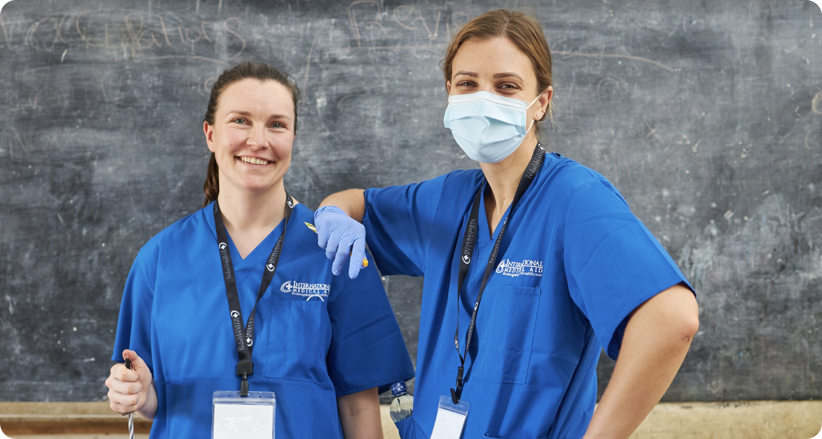 Two individuals in blue medical scrubs, one wearing a mask, smiling in front of a chalkboard.