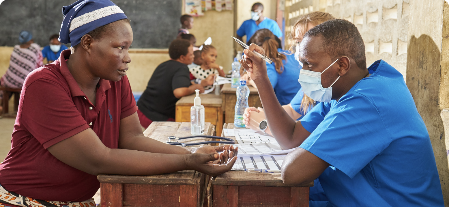 A healthcare professional in blue scrubs examines a person in a community clinic setting, with others in similar attire assisting nearby.
