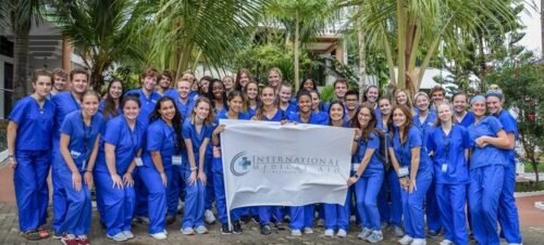 Group of healthcare and pre-med interns in blue scrubs smiling outdoors, holding an International Medical Aid banner in front of palm trees.