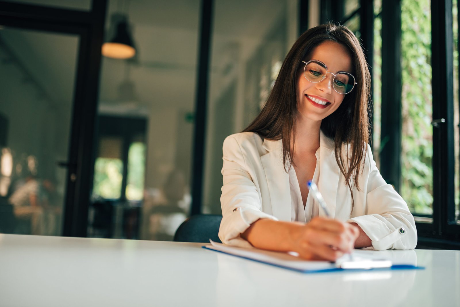 A smiling woman in glasses and a white blazer is writing at a desk, suggesting applying to medical school.