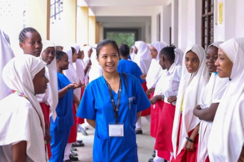 A person in scrubs with a badge interacts with a group of students in uniforms. Keywords: medical internship Kenya.