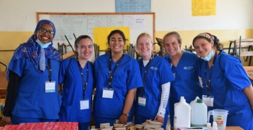 Group of six smiling healthcare interns in blue scrubs standing together in a classroom setting with medical supplies on the table in front of them.