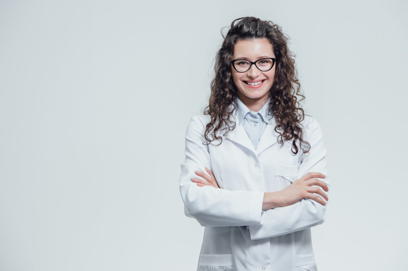A woman with curly hair and glasses, wearing a white lab coat, smiling with arms crossed.