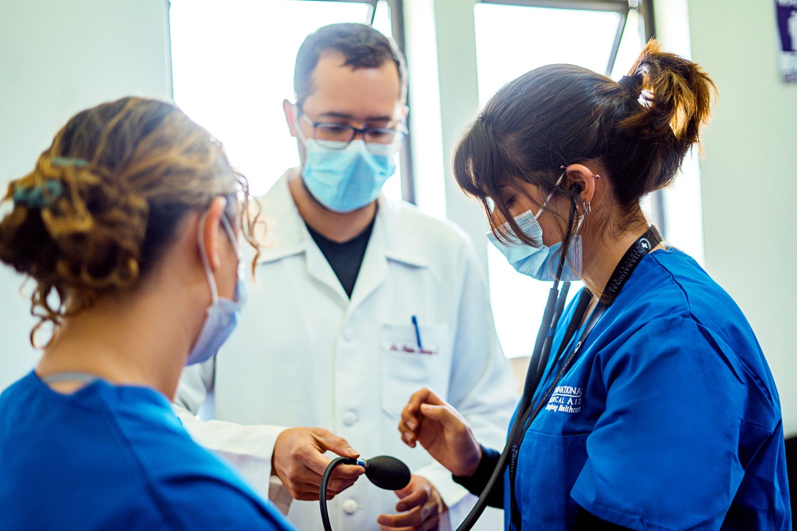 Three healthcare professionals in masks, one in a lab coat and two in blue scrubs, are engaged in medical training, related to internships.