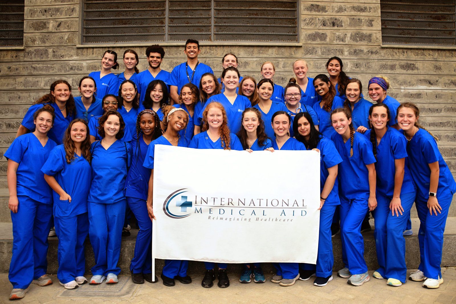 A group of healthcare volunteers in blue scrubs is holding a sign that reads 