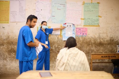 Two people in blue scrubs assist a seated person in a classroom setting, with educational posters on the wall.
