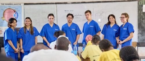 A group of people in blue medical scrubs stand at the front of a classroom, engaging with students, possibly during a medical internship in Kenya.