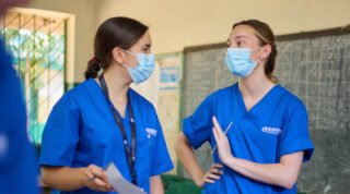 Two people in blue scrubs and masks are conversing in a classroom setting. International medical internship may be relevant.