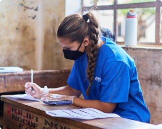 A person in a blue medical uniform and mask writes at a desk, possibly involved in an international medical internship.
