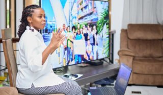 A person is giving a presentation with a laptop and a large screen displaying a group photo in the background.