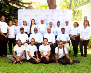 A group of people, some seated and some standing, are posing outside in matching white shirts, likely part of a team or program.