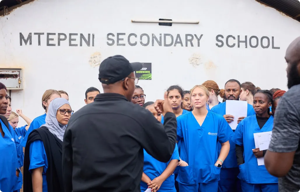 Group of medical interns in blue scrubs receiving instructions from a local guide outside Mtepeni Secondary School.