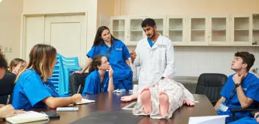 Medical students in blue scrubs attentively watching a clinical instructor demonstrate a procedure on a medical mannequin during a hands-on training session.