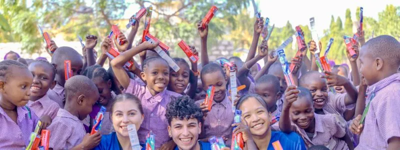 Group of smiling schoolchildren proudly holding up toothbrushes alongside medical interns in blue scrubs.