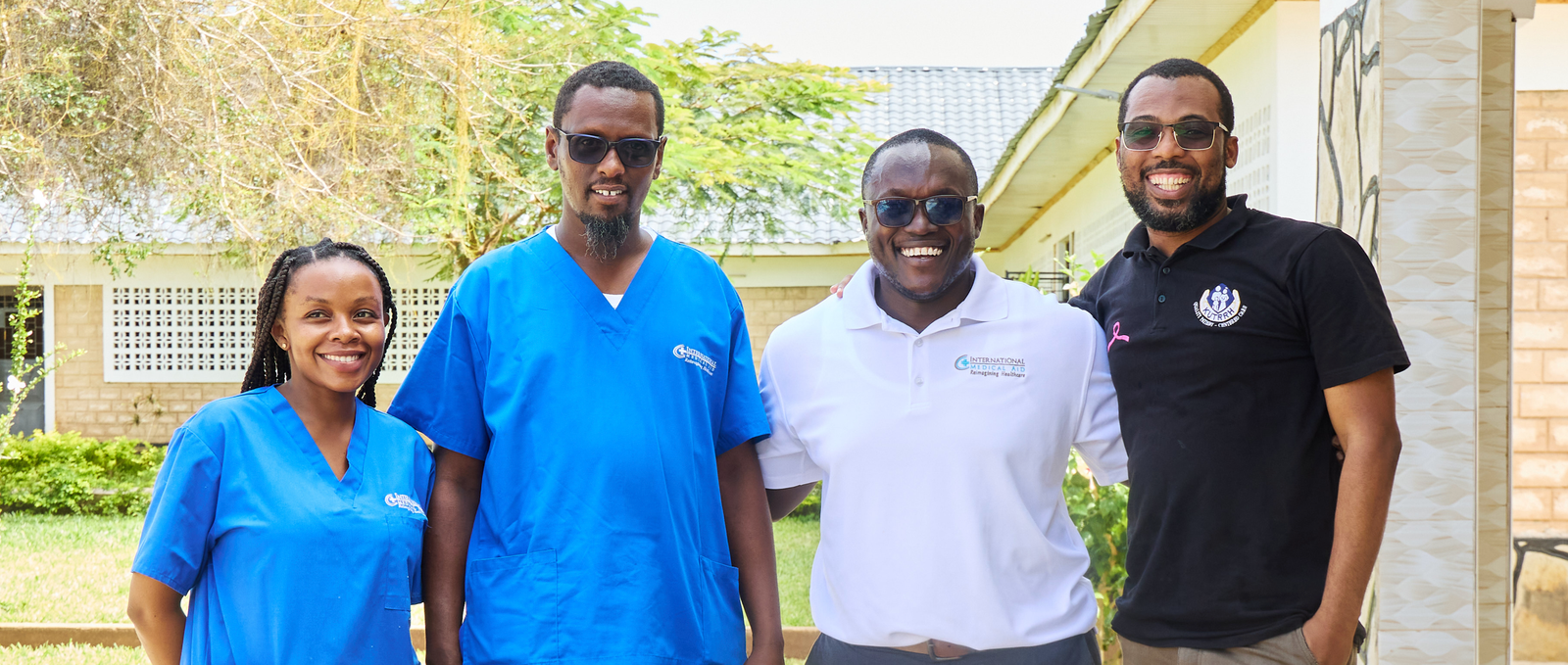 Four people are smiling outside a building, wearing healthcare uniforms and casual attire.