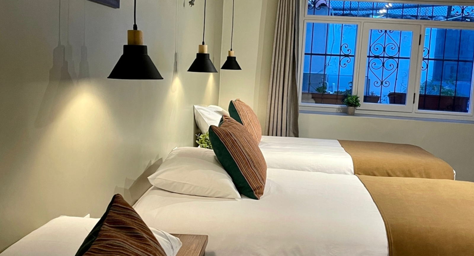 A cozy bedroom with two neatly made beds, pendant lights, and a window with a view.