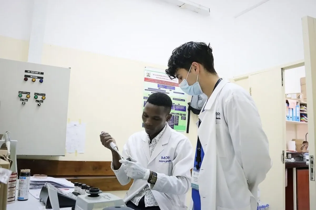Two medical interns in white lab coats working in a clinical laboratory setting, examining a sample using lab equipment.