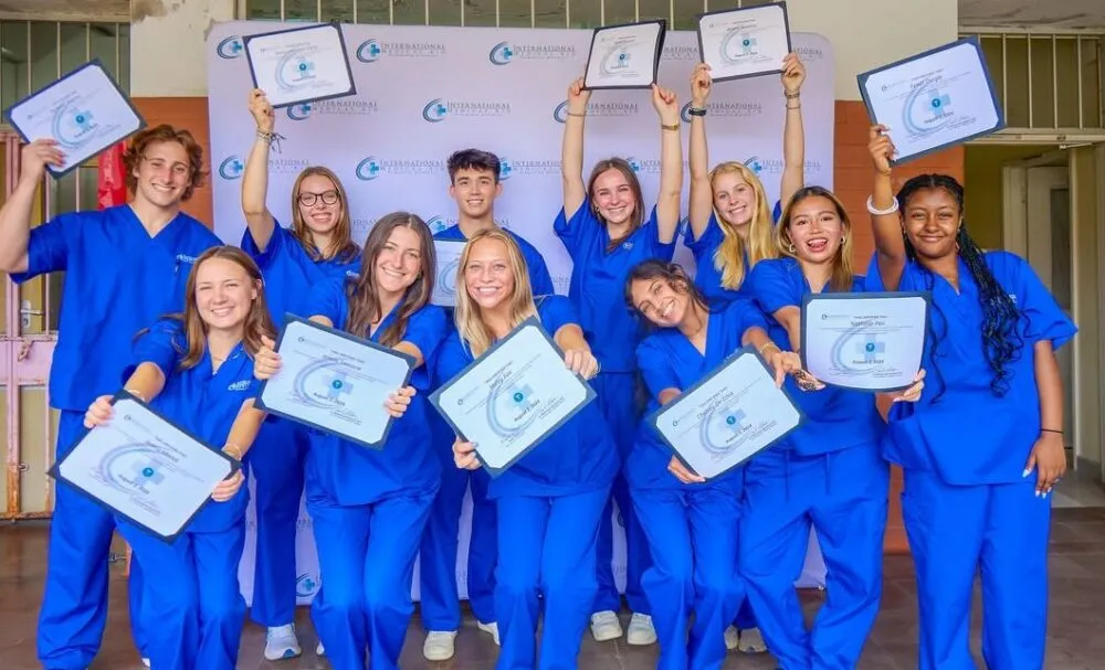 Group of joyful medical interns in blue scrubs proudly holding up certificates in front of a branded backdrop.