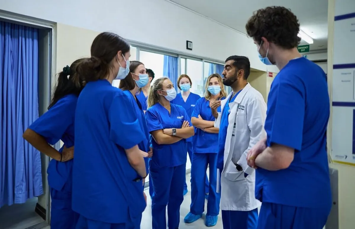 Students in blue scrubs listen to a person in a white coat, likely discussing a medical topic.