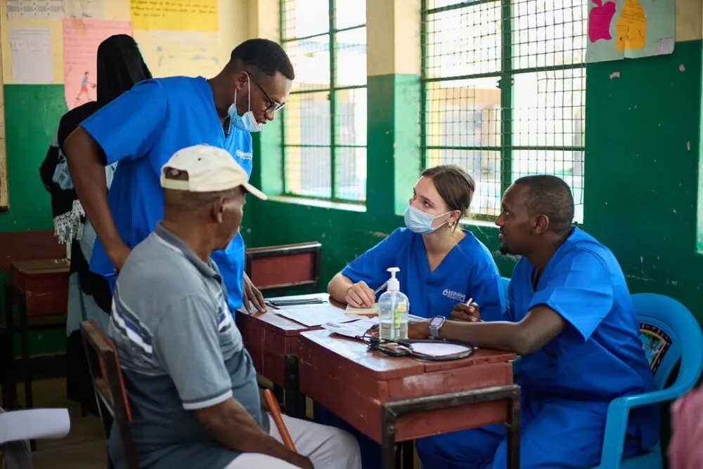 Medical interns in blue scrubs conducting a community health consultation with local residents inside a brightly lit classroom clinic setting.