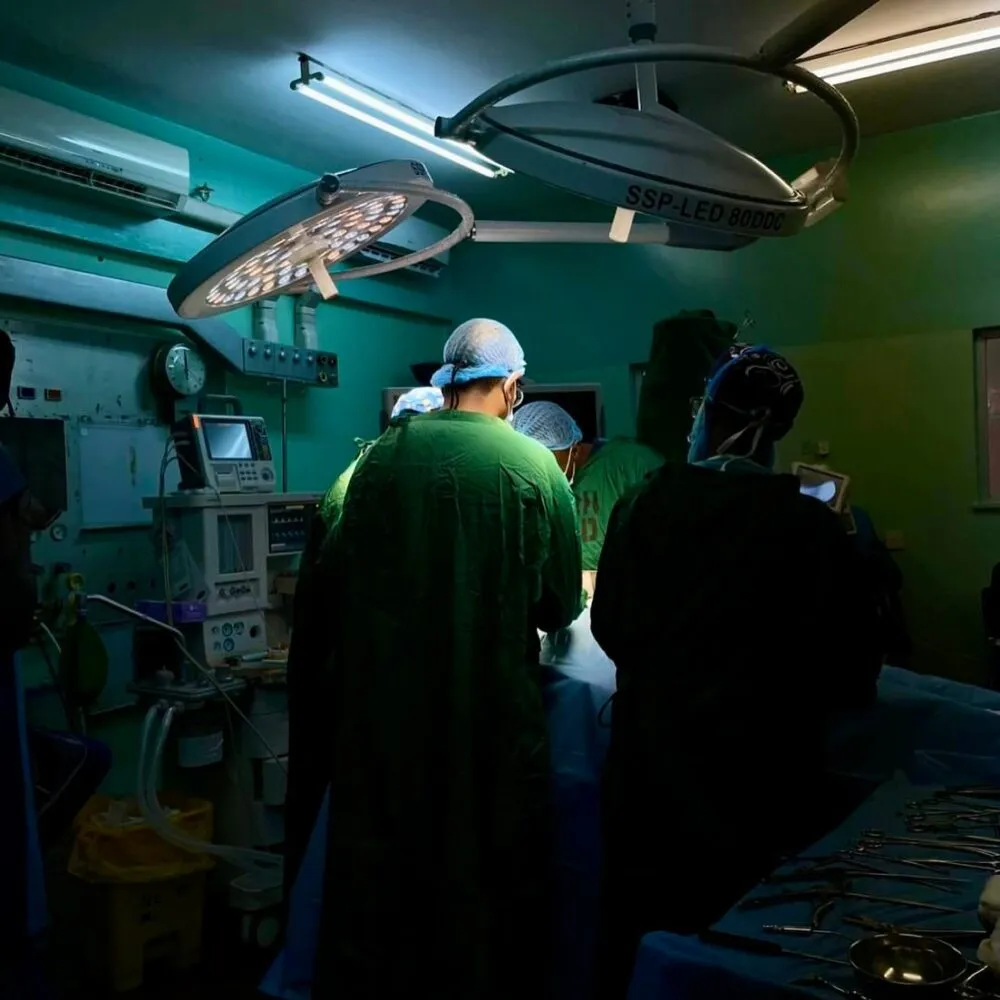 Surgeons in an operating room performing a procedure under bright surgical lights, surrounded by medical equipment in a dimly lit hospital setting.