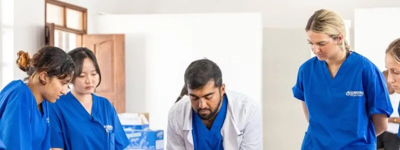 Group of medical interns in blue scrubs observing a clinical demonstration led by a doctor in a white coat inside a bright classroom setting.