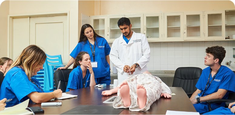 A group of people in medical attire are gathered around a table with a medical dummy, suggesting a healthcare training session.