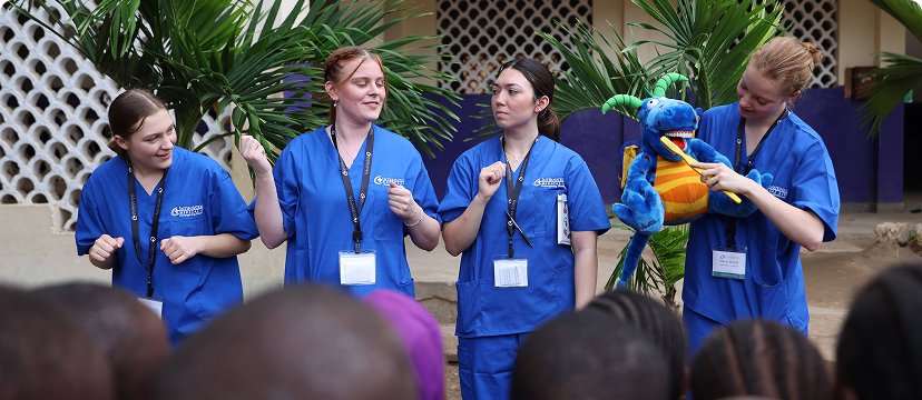 Four people in blue scrubs are engaging with a group, one holding a stuffed dragon toy.