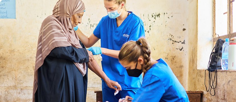 Healthcare workers in blue scrubs are assisting a person in a headscarf within a clinical setting.