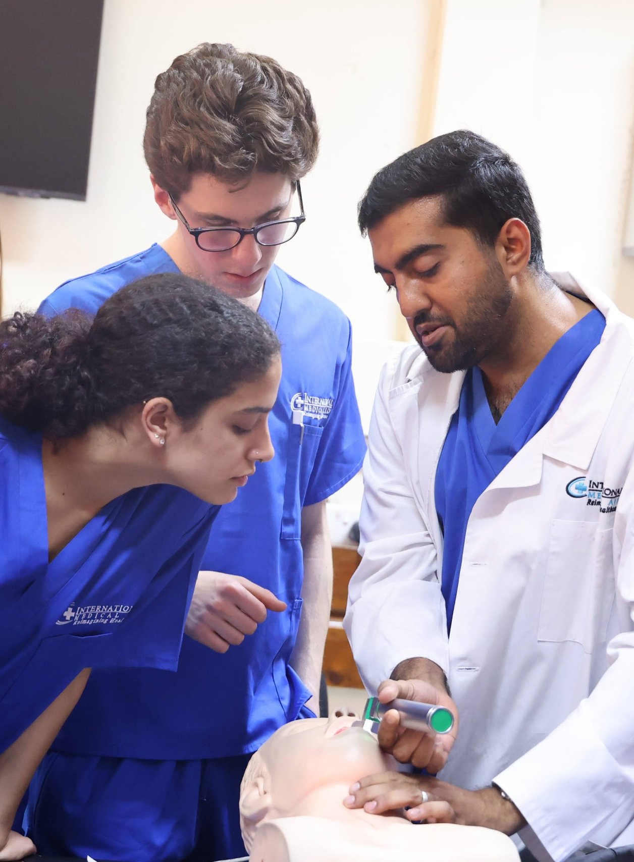 Students in blue scrubs are observing a medical demonstration on a mannequin by a person in a white coat. High school healthcare internships.