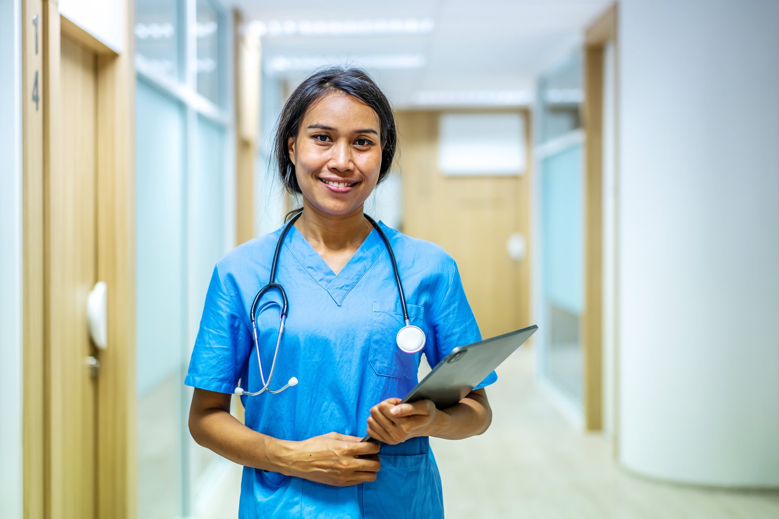 A healthcare professional in scrubs with a stethoscope and clipboard in a medical office hallway.