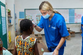 A healthcare worker in blue scrubs examines a child in a classroom setting.