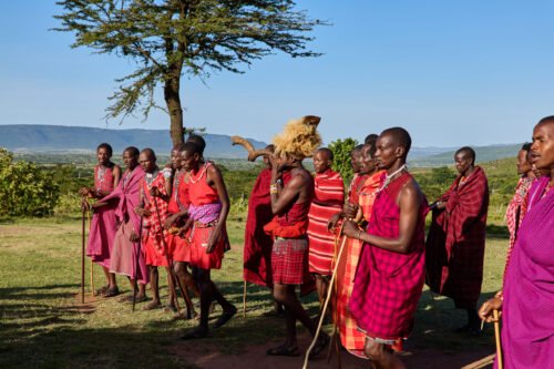A group of people dressed in traditional attire, standing outdoors with a scenic background of trees and hills.