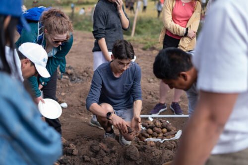 A group of people is gathered around a person examining soil or potatoes in an outdoor setting.