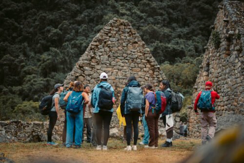 A group of people with backpacks are gathered near ancient stone structures in a forested area.